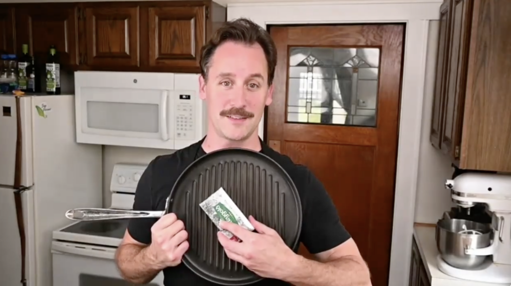 A man with a mustache wearing a black shirt stands in a kitchen, holding up a round black stovetop grill pan. On the surface of the pan, he holds a wrapped stick of butter.